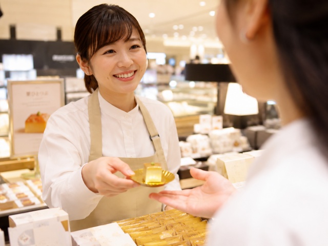 紙製ミニトレーに乗せた試食を笑顔で手渡しする推奨販売スタッフ